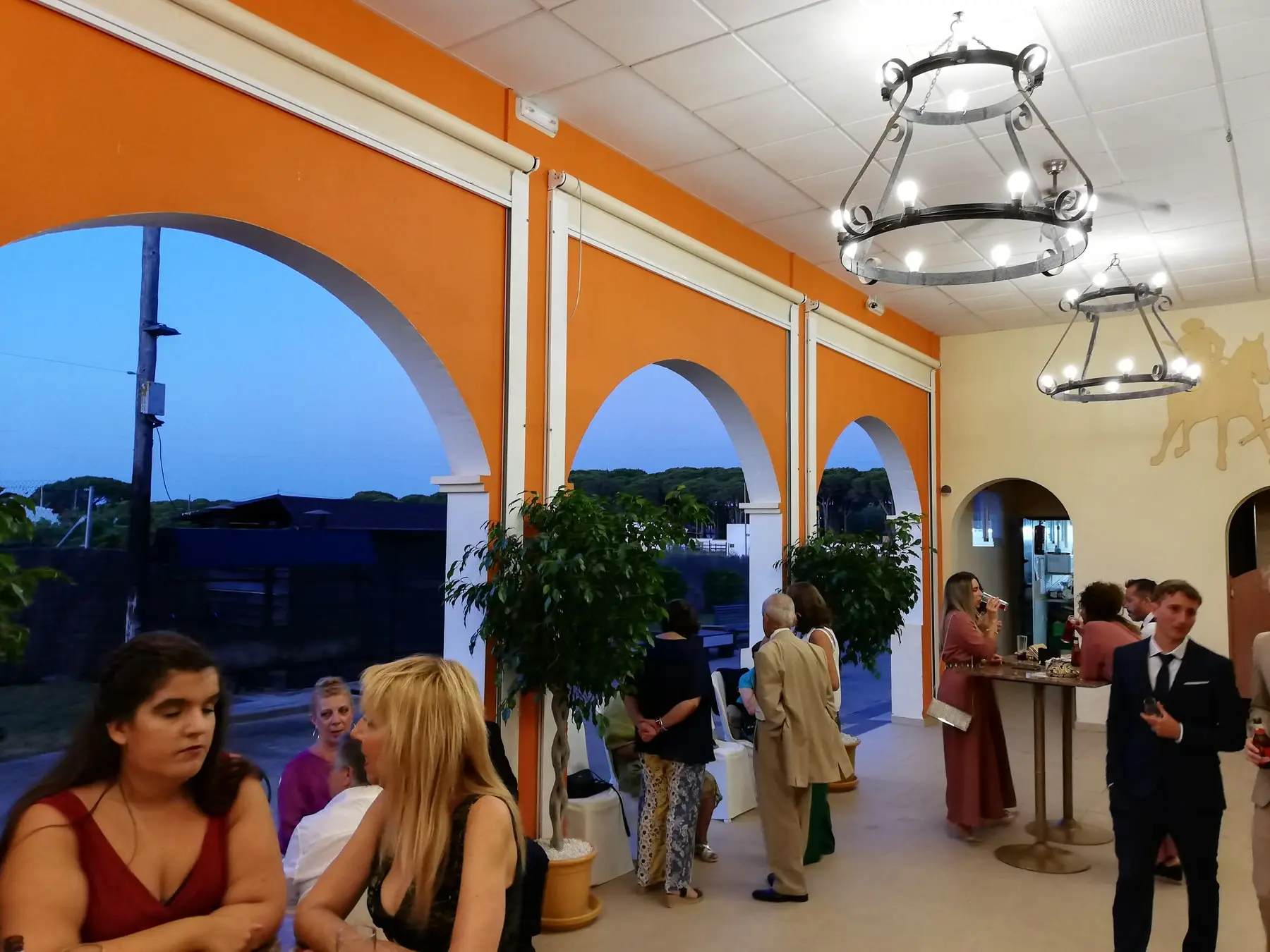 Invitados charlando bajo los arcos de la galería de paredes naranjas, con lámparas de forja y pinos al fondo