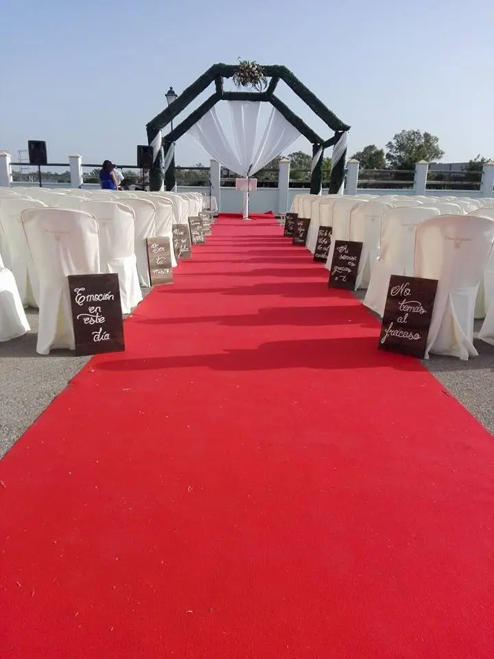 Una alfombra roja entre filas de sillas blancas hasta un arco de ceremonia con tela blanca, con letreros de madera con frases