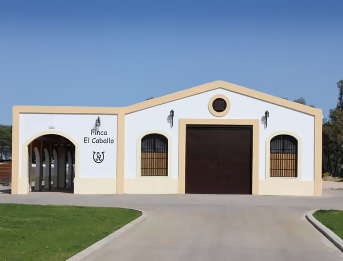 La fachada blanca de Finca El Caballo con su arco, el nombre y la herradura sobre la pared, una puerta de madera oscura y ventanas con reja